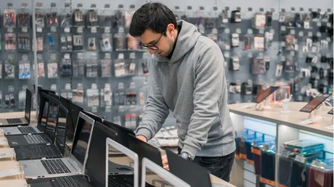 Bir erkek, teknoloji mağazasında dizüstü bilgisayarlara bakıyor. Arka planda telefon aksesuarları sergileniyor.