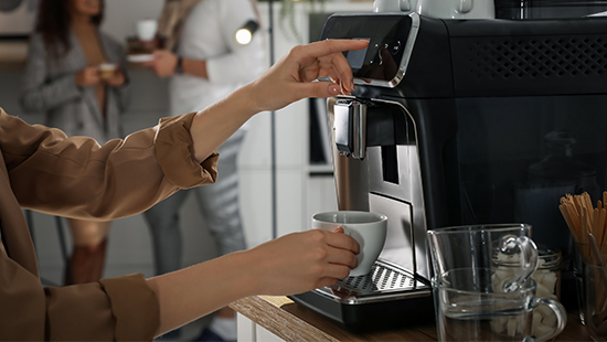 Bir kadın, kahve makinesinden bardağa kahve dolduruyor. Arka planda bulanık insanlar var.