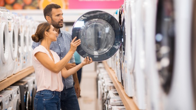 Bir çift, beyaz eşya mağazasında bir çamaşır makinesinin kapağını inceliyor. Raflarda sıralanmış makineler görülüyor.