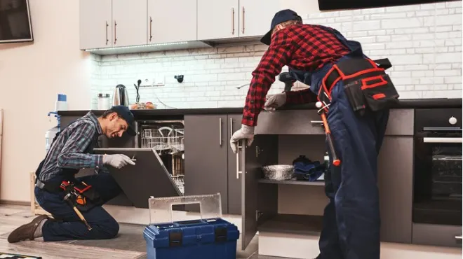 Mutfakta, iki tamirci dolapları tamir ediyor. Biri diz çökmüş, diğeri ayakta. Alet kutusu yerde.