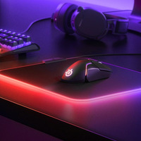 Siyah mouse, kırmızı kenarlı mouse pad, klavye ve kulaklık mor ışık altında.
