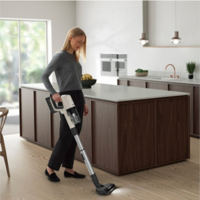 Sarışın kadın mutfakta elektrikli süpürge kullanıyor. Arka planda ada tezgahı ve dolaplar var.