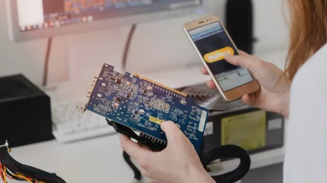 Mavi devre kartı tutan ve telefon kullanan kişi. Arka planda monitör, klavye ve bilgisayar kasası var.