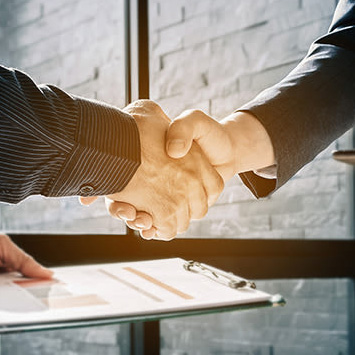 İki kişi, masa üzerinde belgelerle tokalaşıyor. Arka planda tuğla desenli duvar var.