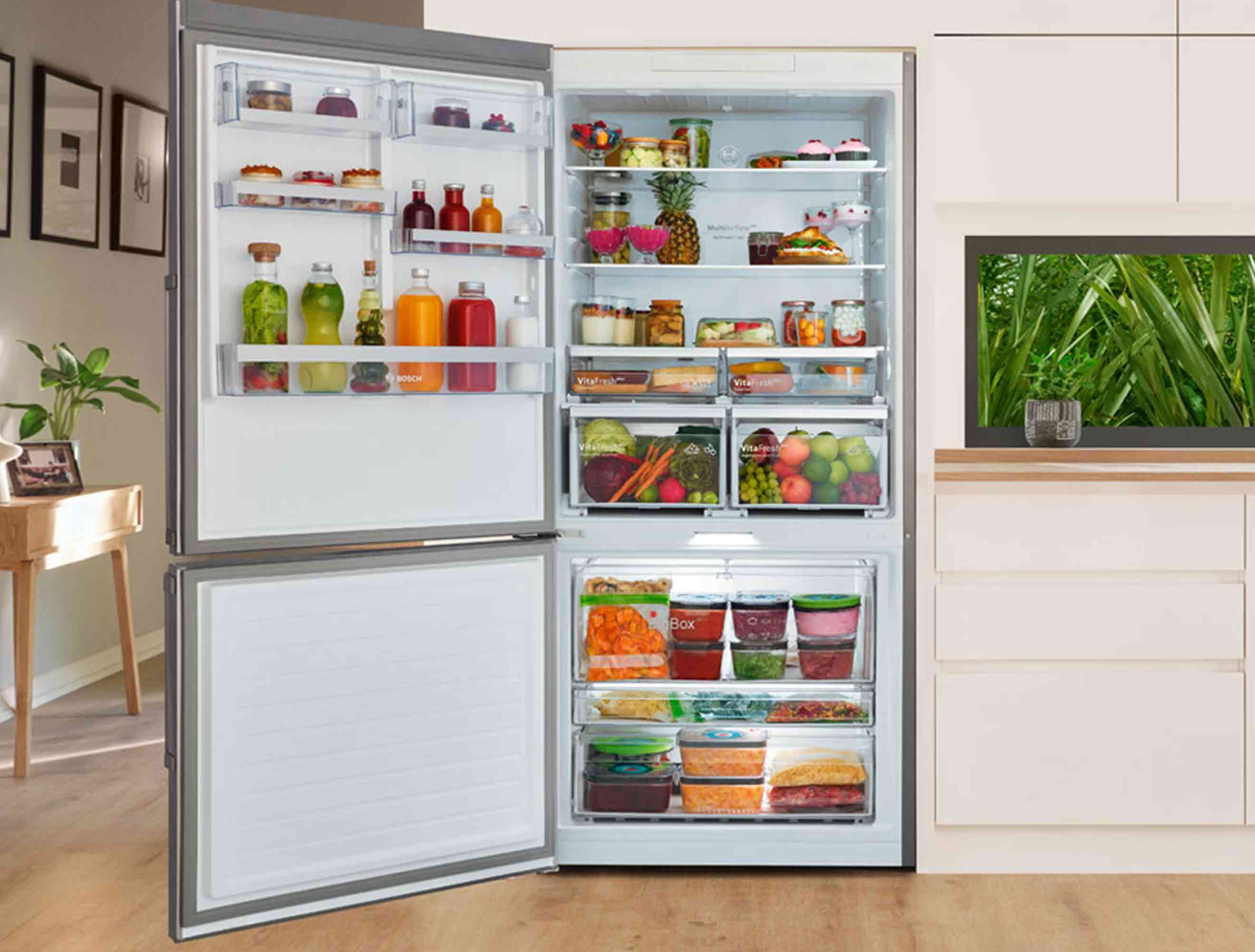 Açık kapılı, gümüş çerçeveli buzdolabı, meyve, sebze ve içeceklerle dolu. Mutfak dolapları ve pencere arka planda.