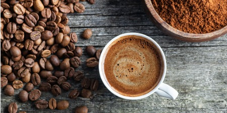 Ahşap zeminde kahve çekirdekleri, bir fincan kahve ve bir kasede öğütülmüş kahve bulunmaktadır.