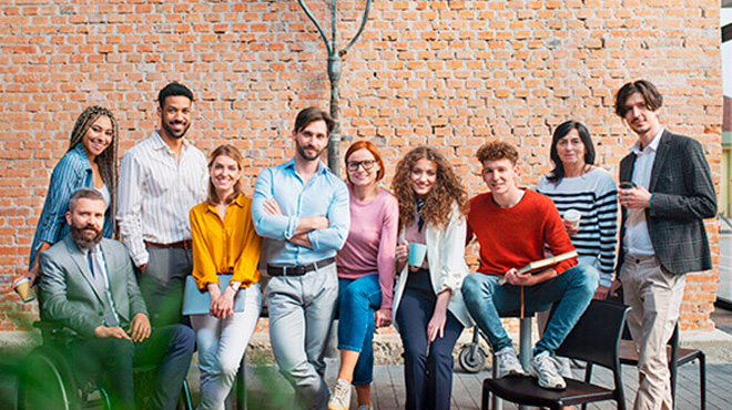 Tuhaf giyimli, çeşitli insanlar tuğla duvar önünde poz veriyor. Bir kişi tekerlekli sandalyede oturuyor.