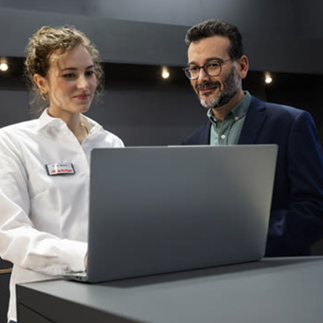 Bir kadın ve bir erkek, gri bir dizüstü bilgisayara bakıyor. Kadın beyaz gömlek giyiyor, erkek ise lacivert bir ceket giyiyor ve gözlük takıyor.