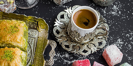 Siyah zeminde gümüş tepside baklava, kahve fincanı ve lokumlar görülüyor.