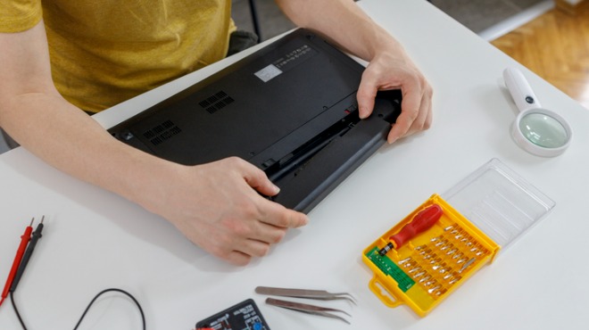 Bir kişi, sarı bir gömlek giymiş, siyah bir dizüstü bilgisayarın alt kapağını açıyor. Aletler ve büyüteç masada.