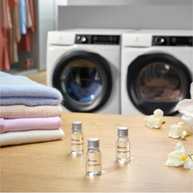 Çamaşır makinelerinin önünde, şişeler, çiçekler ve katlanmış giysiler bulunan ahşap bir yüzey.