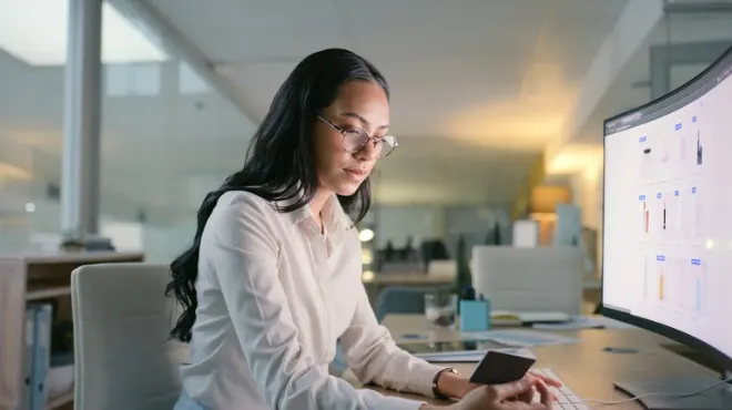 Ofiste, beyaz gömlekli, gözlüklü kadın ekrana bakıp kart tutuyor. Arka planda ofis ortamı var.