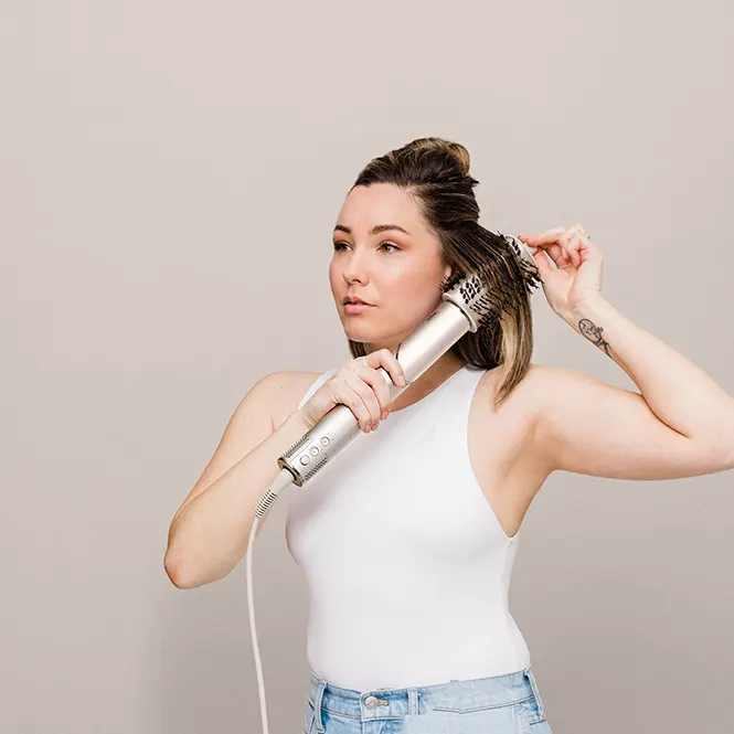 Beyaz atletli kadın, saçını elektrikli fırçayla şekillendiriyor. Fırça gümüş renginde, duvar pembe.