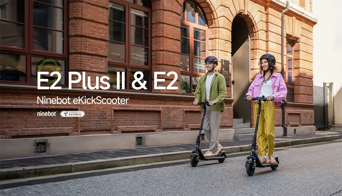 İki kişi Ninebot eKickScooter'lar üzerinde, tuğla binanın önünde duruyor. Erkek yeşil, kadın pembe ceket giyiyor.