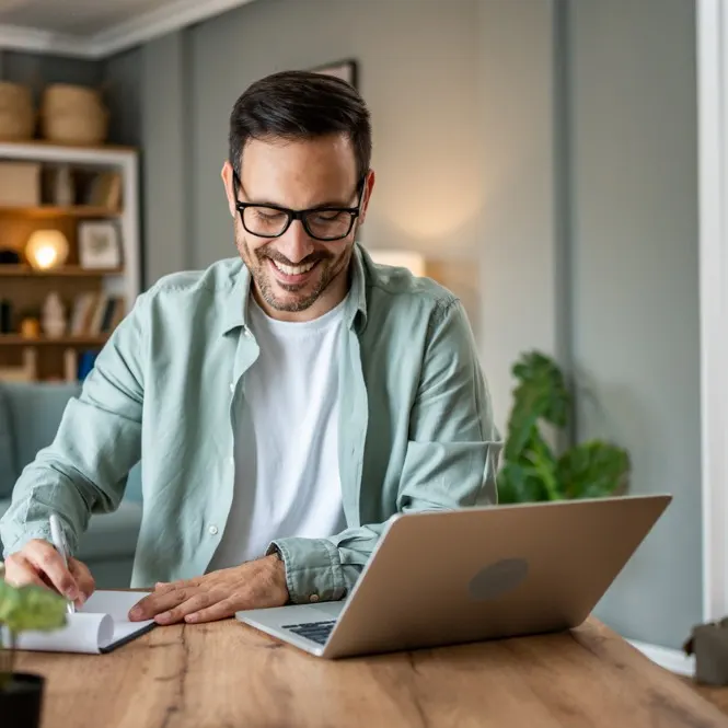 Gözlüklü adam ahşap masada dizüstü bilgisayar kullanıyor, not alıyor. Arka planda raf ve bitki var.
