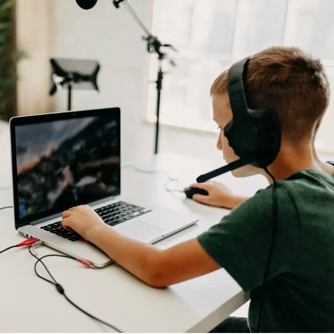 Oturma odasında kulaklıklı, dizüstü bilgisayarda oyun oynayan veya ders çalışan çocuk. Beyaz masada mikrofon ve kulaklık var.