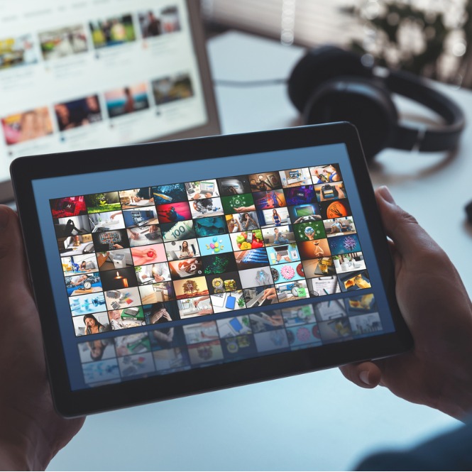 Tablet tutan ellerde, ekranda çok sayıda küçük resim var. Arka planda kulaklık ve başka bir ekran görülüyor.