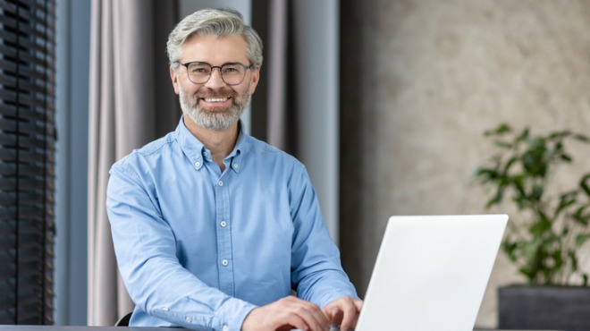 Gülümseyen, sakallı, gözlüklü adam mavi gömlekle dizüstü bilgisayar kullanıyor. Arka planda bitki ve perde var.