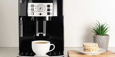 Siyah kahve makinesi, beyaz fincan, yiyecek ve saksı bitkisiyle birlikte açık renkli bir tezgahta duruyor.