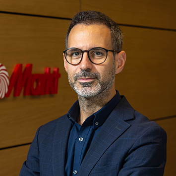 Gözlüklü, sakallı, lacivert ceketli bir adam, kahverengi zemin önünde duruyor. Arka planda kırmızı bir logo var.