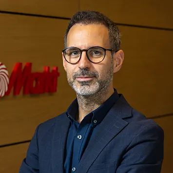 Gözlüklü, sakallı, lacivert ceketli bir adam, kahverengi zemin önünde duruyor. Arka planda kırmızı bir logo var.