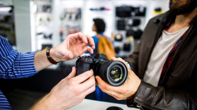 Bir adam bir kamerayı inceliyor. Diğer adam mavi çizgili bir gömlek giyiyor. Arka planda raflar var.