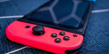 Kırmızı ve mavi Joy-Con'lu Nintendo Switch, mavi bir yüzey üzerinde duruyor. Oyun konsolunun ekranı ve kontrol düğmeleri görülüyor.