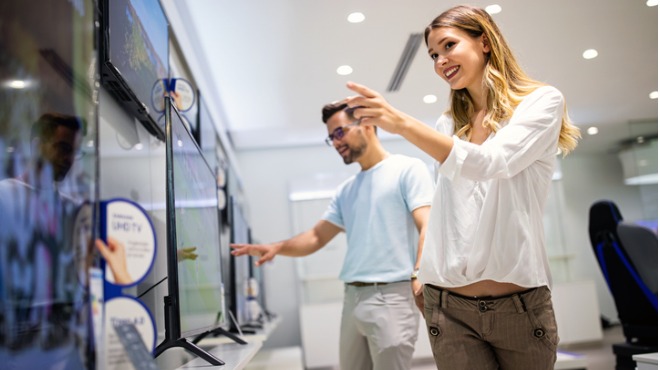 Bir TV mağazasında TV'lere bakan, ellerini uzatan bir kadın ve bir adam.