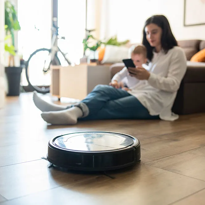 Oturma odasında, annenin kucağında bebekle telefon kullandığı, yerde robot süpürgenin çalıştığı bir sahne.