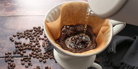 Beyaz bir demlikten kahve filtresine su dökülüyor. Kahve çekirdekleri ve koyu renkli bir bez de görülebilir.