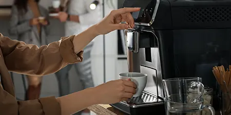 Kahve makinesi kullanan kişi, beyaz fincanı dolduruyor. Arka planda diğer insanlar ve mutfak tezgahı var.