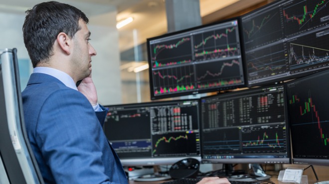 Mavi takım elbiseli adam telefonla konuşuyor, önünde borsa grafiklerinin olduğu çoklu ekranlar var.