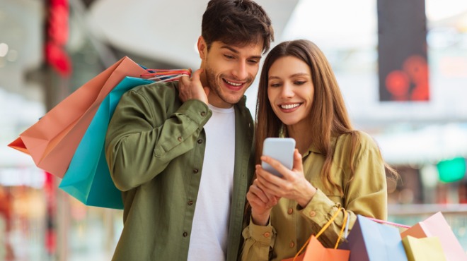 Gülümseyen bir çift alışveriş merkezinde, akıllı telefona bakıyorlar. Adamın omzunda renkli alışveriş çantaları var.
