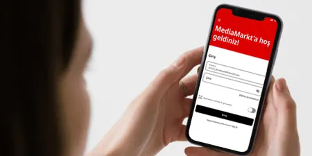 Bir kişi, MediaMarkt uygulamasının açık olduğu bir akıllı telefon tutuyor. Ekran kırmızı ve beyaz renkte.