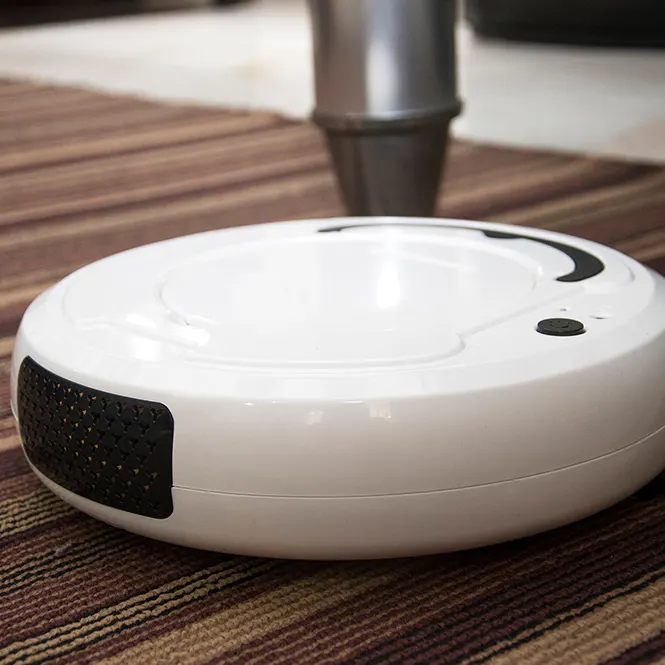 Oda üzerinde halı üzerinde duran beyaz robot süpürge ve arka planda gümüş rengi masa ayağı görülmektedir.