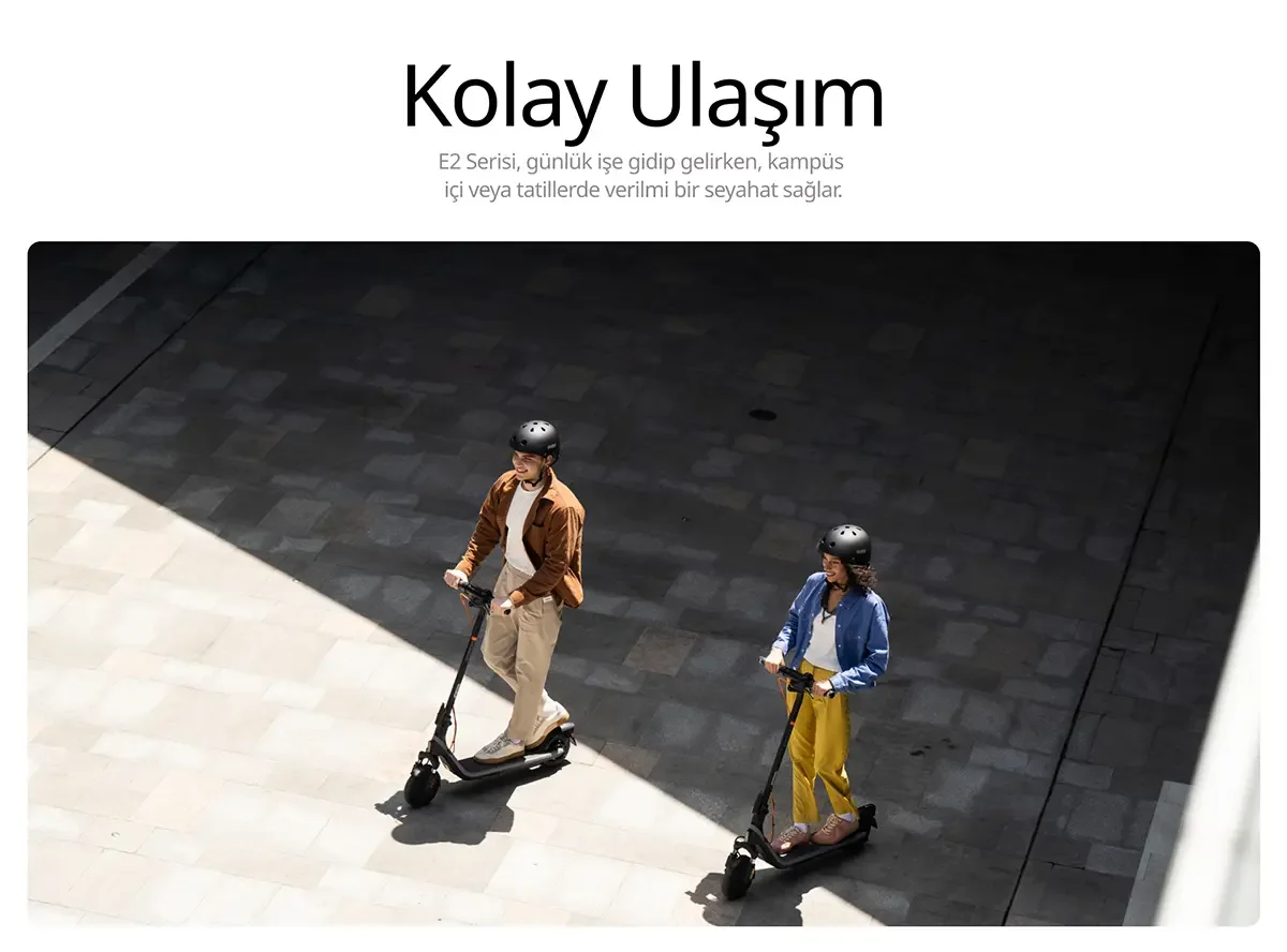 İki kişi kask takmış, elektrikli scooter sürüyor. Erkek kahverengi ceketli, kadın sarı pantolonlu. Zemin taş döşeli.