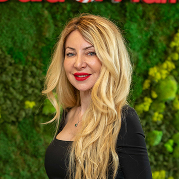 Siyah bluzlu, sarı saçlı kadın, yeşil bitki duvarı önünde kırmızı rujlu gülümsüyor.