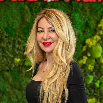 Siyah bluzlu, sarı saçlı kadın, yeşil bitki duvarı önünde kırmızı rujlu gülümsüyor.