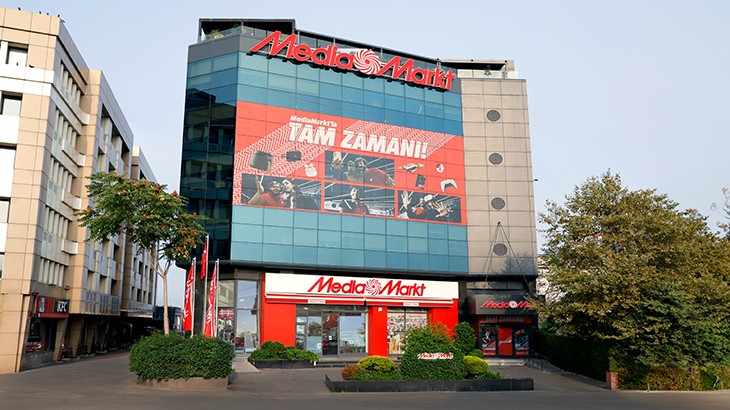 Dışarıdan bir Media Markt mağazası. Üzerinde kırmızı bir logo ve "Tam Zamanı!" yazan büyük bir reklam panosu var.