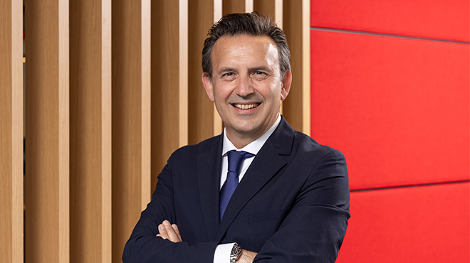 Ofiste oturan, gri ceketli, beyaz gömlekli, kahve kupası tutan, gülen bir adam. Arka planda pencere ve bilgisayar var.