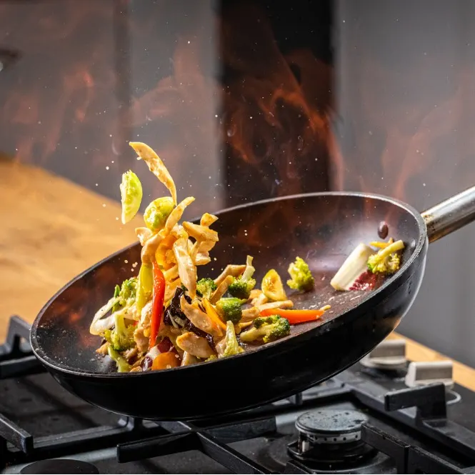Ocakta wok tava içinde sebzeler ve et soteleniyor. Alevler ve hareketli malzemeler yemek pişirme anını vurguluyor.