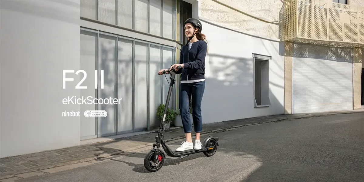 A woman wearing a helmet is traveling around the city on a Ninebot F2 II eKickScooter. A building and a garage door are in the background.
