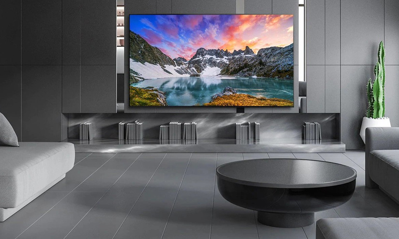 Geniş ekran TV'nin altında kitaplar olan, gri tonlamalı modern bir oturma odası görülmektedir.