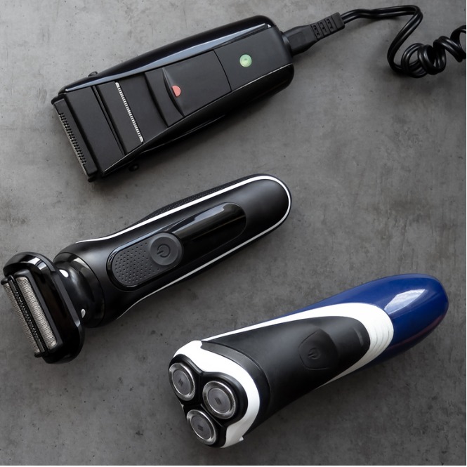 Siyah, beyaz ve mavi tıraş makineleri ve şarj cihazı gri bir yüzeyde duruyor.