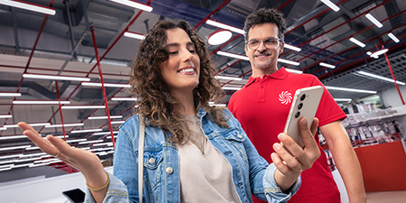 Bir kadın cep telefonu tutuyor, gülümsüyor. Arka planda kırmızı tişörtlü bir adam var.