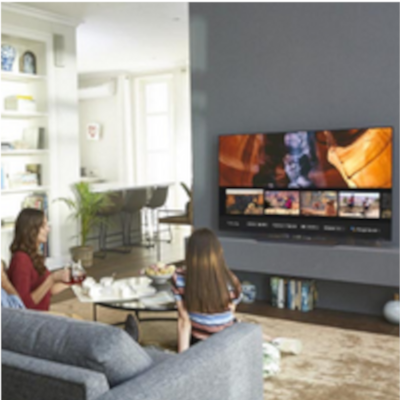 Oturma odasında TV izleyen aile. Gri kanepe, sehpa ve duvarda büyük ekran TV. Beyaz raf ünitesi ve halı zemin.