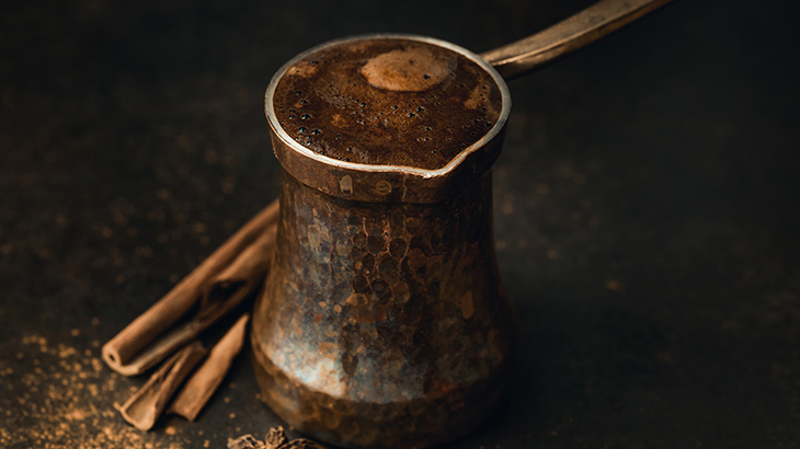 Koyu zeminde cezve, kahve ve tarçın çubukları görülüyor. Cezve bakır renginde, kahve köpüklü.