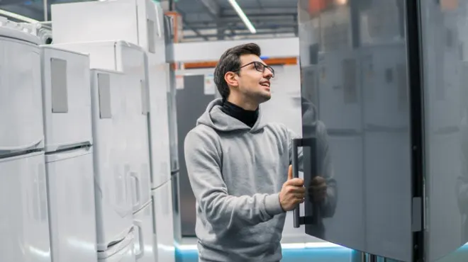 Gri kapüşonlu, gözlüklü bir adam buzdolabı inceliyor. Arkada beyaz eşyalar var.