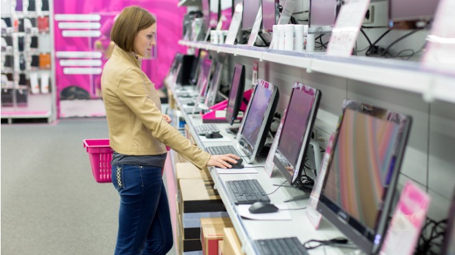 Bir kadın elektronik eşya mağazasında bilgisayarları inceliyor. Pembe bir alışveriş sepeti taşıyor.