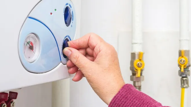 Beyaz duvara monte edilmiş, mavi detaylı bir kombinin düğmesini çeviren bir el. Arka planda sarı vanalı borular var.